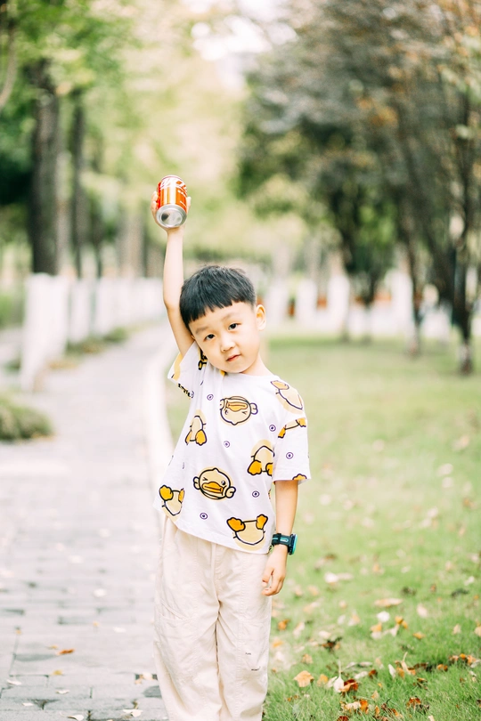 一个穿着睡衣的小男孩站在树叶覆盖的人行道上 手里拿着一个小罐子 看着镜头