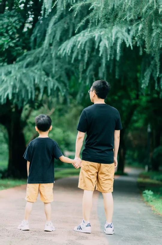 一个小男孩和一个年轻男子手拉手在公园小径上走