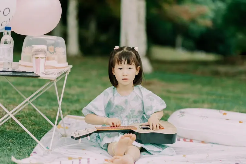 一个穿着连衣裙的小女孩坐在毛毯上 在公园里的野餐上阅读一本书 周围有粉红色的气球。