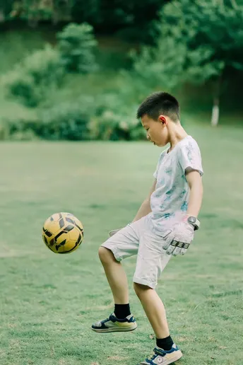 一个年轻的男孩在草地上踢足球 手里拿着一个黄色的球和手套。