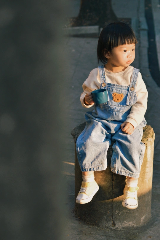 一个小女孩穿着工作服坐在水泥长凳上 手里拿着一杯东西。