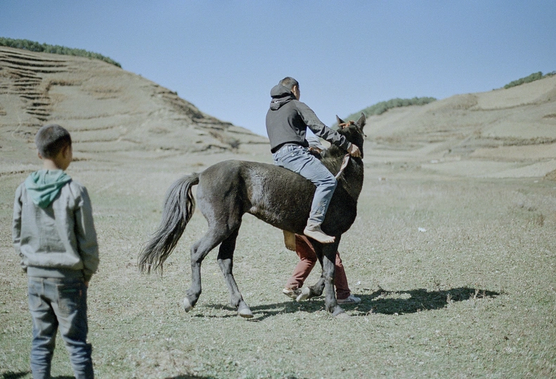 一个年轻男孩骑着一匹马在沙漠中 背景是起伏的山丘 一个男人在观看。