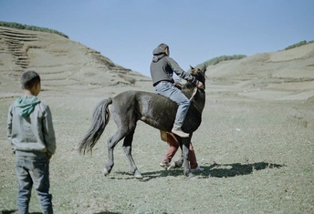 一个年轻男孩骑着一匹马在沙漠中 背景是起伏的山丘 一个男人在观看。
