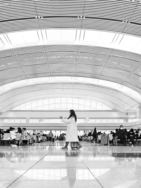 一张黑白照片 机场航站楼 有人走动 一位穿大衣的女人站在行李旁