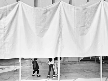 一些年轻人站在一顶白色的帐篷下 一个男孩在建筑物前骑滑板。