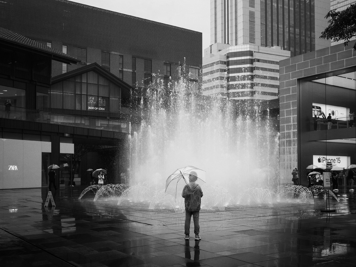 一张黑白照片 一位男子站在城市中的喷泉前拿着一把伞。