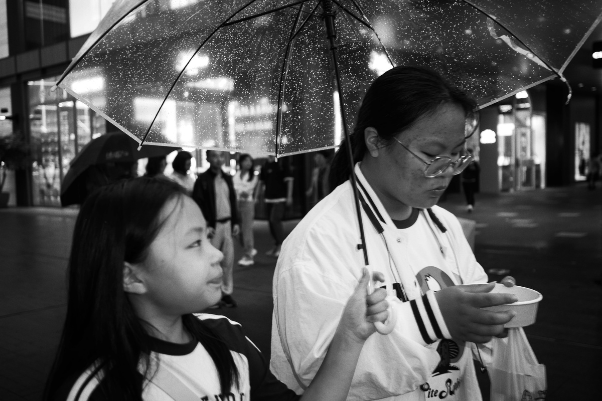 一个拿着黑白色雨伞站在小女孩旁边 小女孩正在雨中用碗吃饭的繁忙街道上。