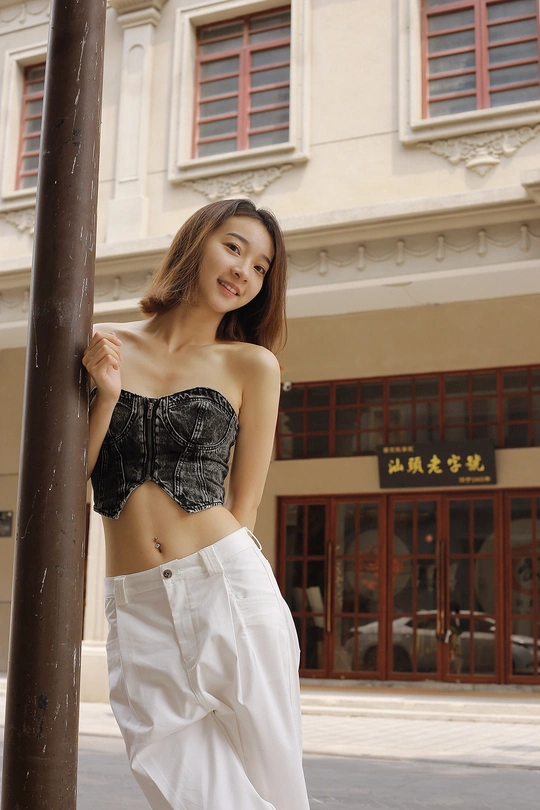 一位穿着黑色上衣和白色裤子的年轻女子 靠在一根街边的柱子上 前面是一座建筑。