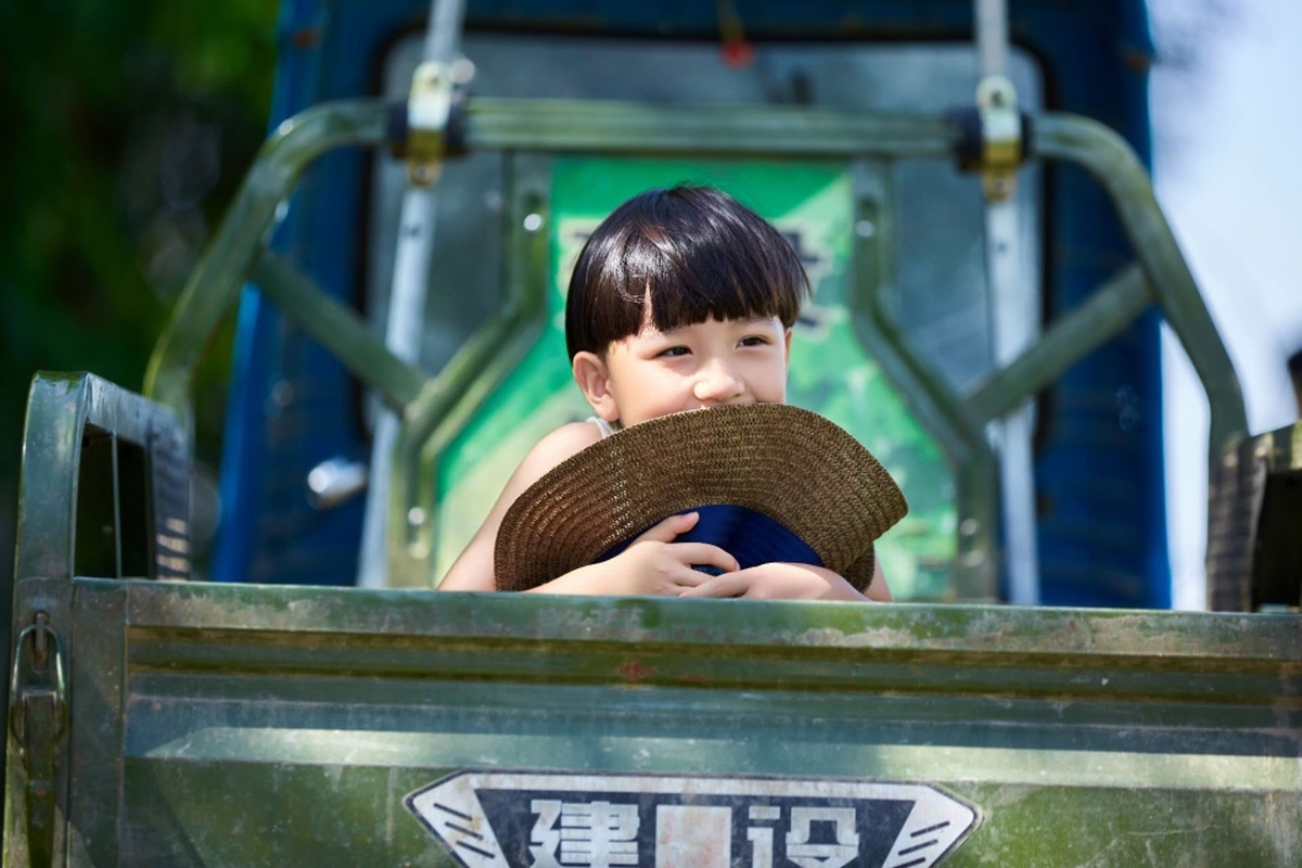 一个年轻男孩戴着帽子坐在一辆小卡车里 另一个小男孩在旁边看着。