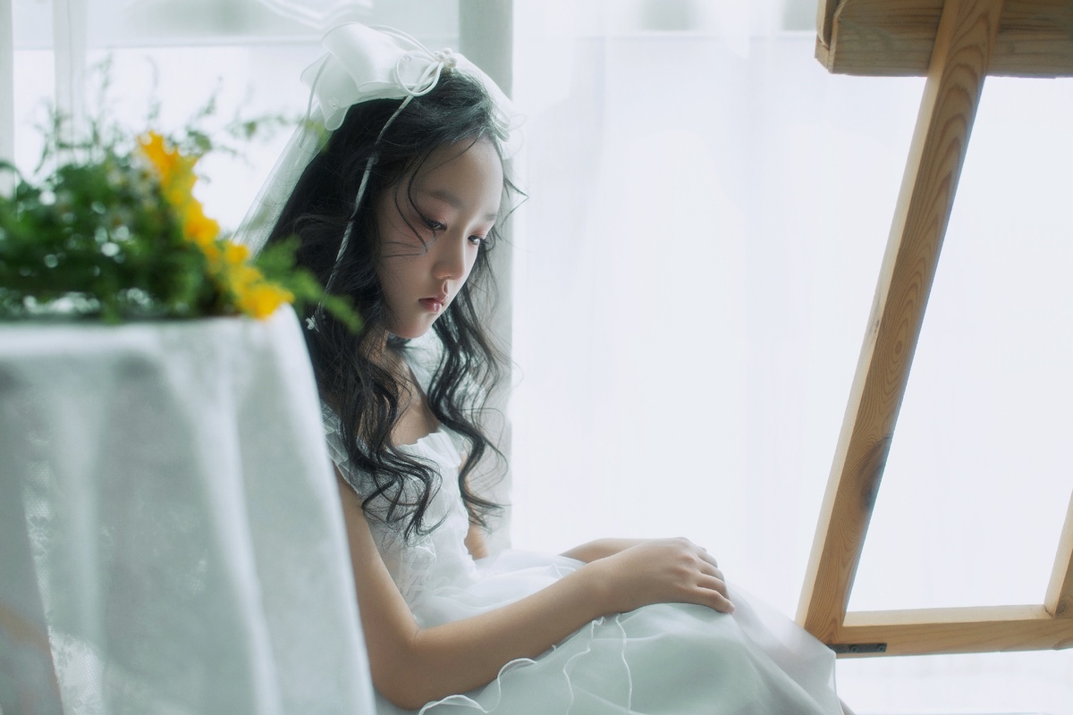 一个小女孩穿着白色连衣裙坐在椅子上 面前有一扇窗户 桌子上开着花。
