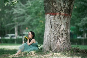 一位穿着绿色连衣裙的年轻女子坐在草地上 靠在一棵树旁 在公园里。