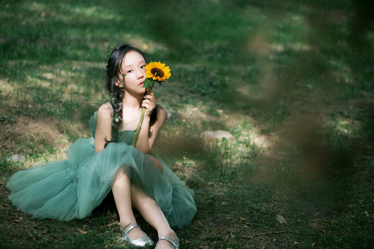 一个小女孩穿着绿色裙子坐在森林地上 手里拿着一朵花。
