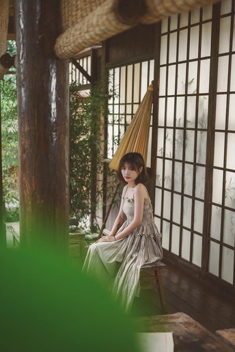 一个穿着白色连衣裙的年轻女子坐在带有吊床和窗户的房间里。