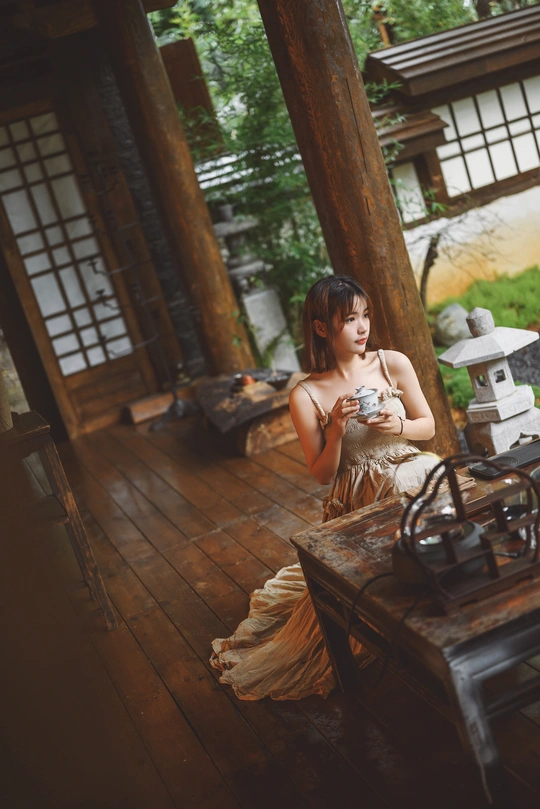 一位穿着连衣裙的年轻女子手持相机坐在木制桌子前 房子就在她身后。
