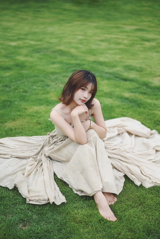 一个美丽的年轻女子穿着一件白色连衣裙坐在绿色的草地上的一块毛毯上