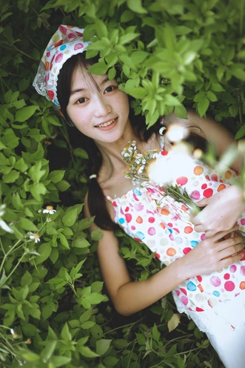 一位穿着连衣裙和帽子 手里拿着一朵小花的年轻女子 站在一片绿色植物和灌木丛的田野中。