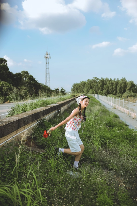 一位穿着连衣裙的年轻女子手里拿着一朵花 沿着围着高草的围墙走。