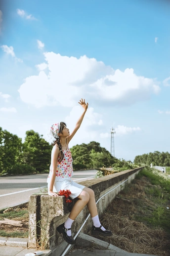一个年轻女子坐在靠近公路的墙边 手里拿着花 正在仰望天空。