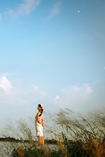 一个穿着白色连衣裙的年轻女子站在一片高草丛生的草地附近的水体旁边。