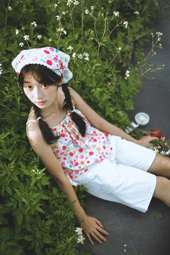 一个穿着裙子和帽子的小女孩坐在草地上 旁边有一丛花。