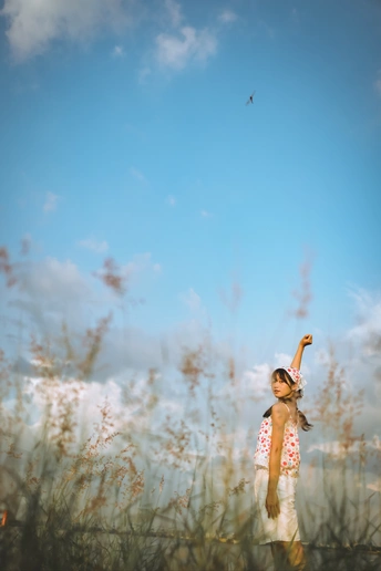 一个小女孩站在一片高草地上 伸开双臂 天空中一只风筝在飞翔。