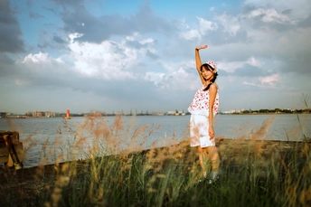 一个穿着连衣裙的小女孩站在海滩上 举起双臂 手指指向水面 一只风筝在天空中飞翔。