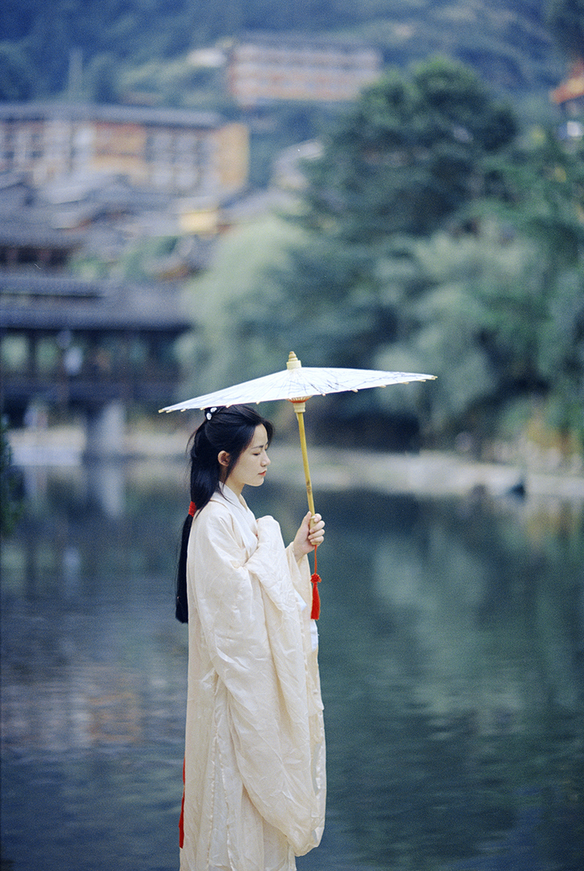 一位穿着白色连衣裙 手持雨伞遮阳的女士站在水边。