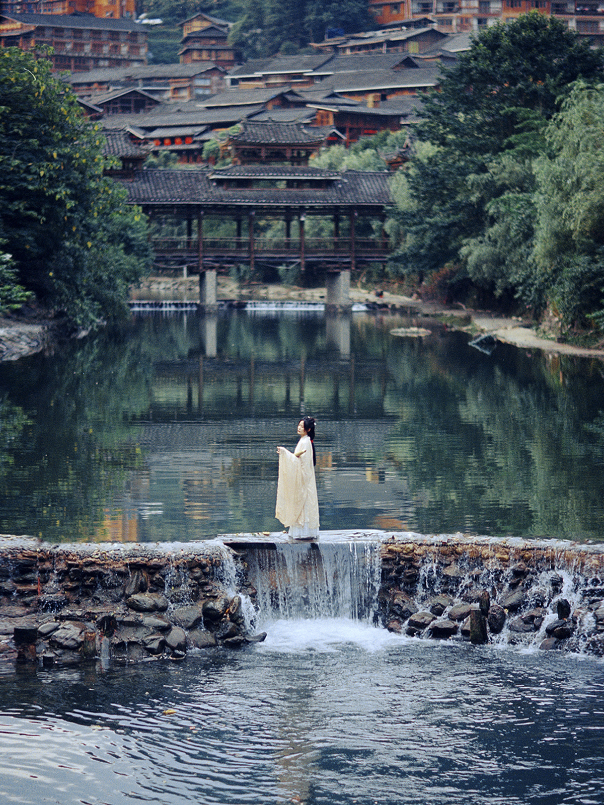 一个穿白色连衣裙的女人站在一座跨水桥上 手里拿着一把伞。