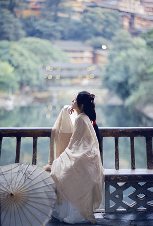 一位穿着白色连衣裙 手持打开的雨伞 坐在阳台 overlooking 水面上的女士。