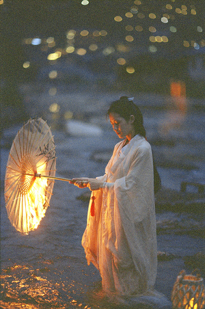 一个穿着白色连衣裙的年轻女子手握一盏灯伞站在夜晚的海滩上。