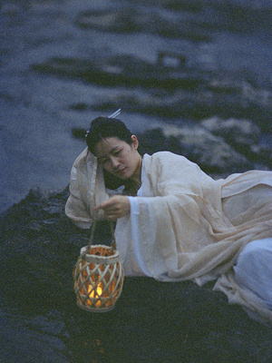 一位身着白色长袍的女性 手持点燃的蜡烛坐在河流中央的一块岩石上 而水中有一位男性。