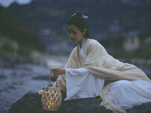 一个穿着白色长袍的年轻女子拿着一个灯笼 里面有一支蜡烛 坐在靠近水边的一块石头上。