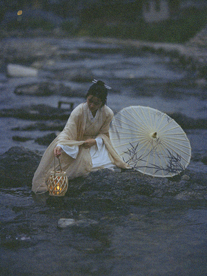 一个穿着白色连衣裙的女人拿着扇子和雨伞坐在靠水的岩石上