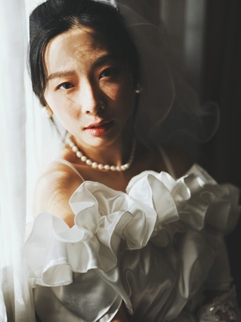 一个戴白色头纱的女士站在窗前 窗帘是透明的。