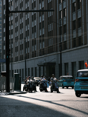 几个人骑着摩托车在一条有高楼大厦和一辆蓝色汽车的城市街道上行驶