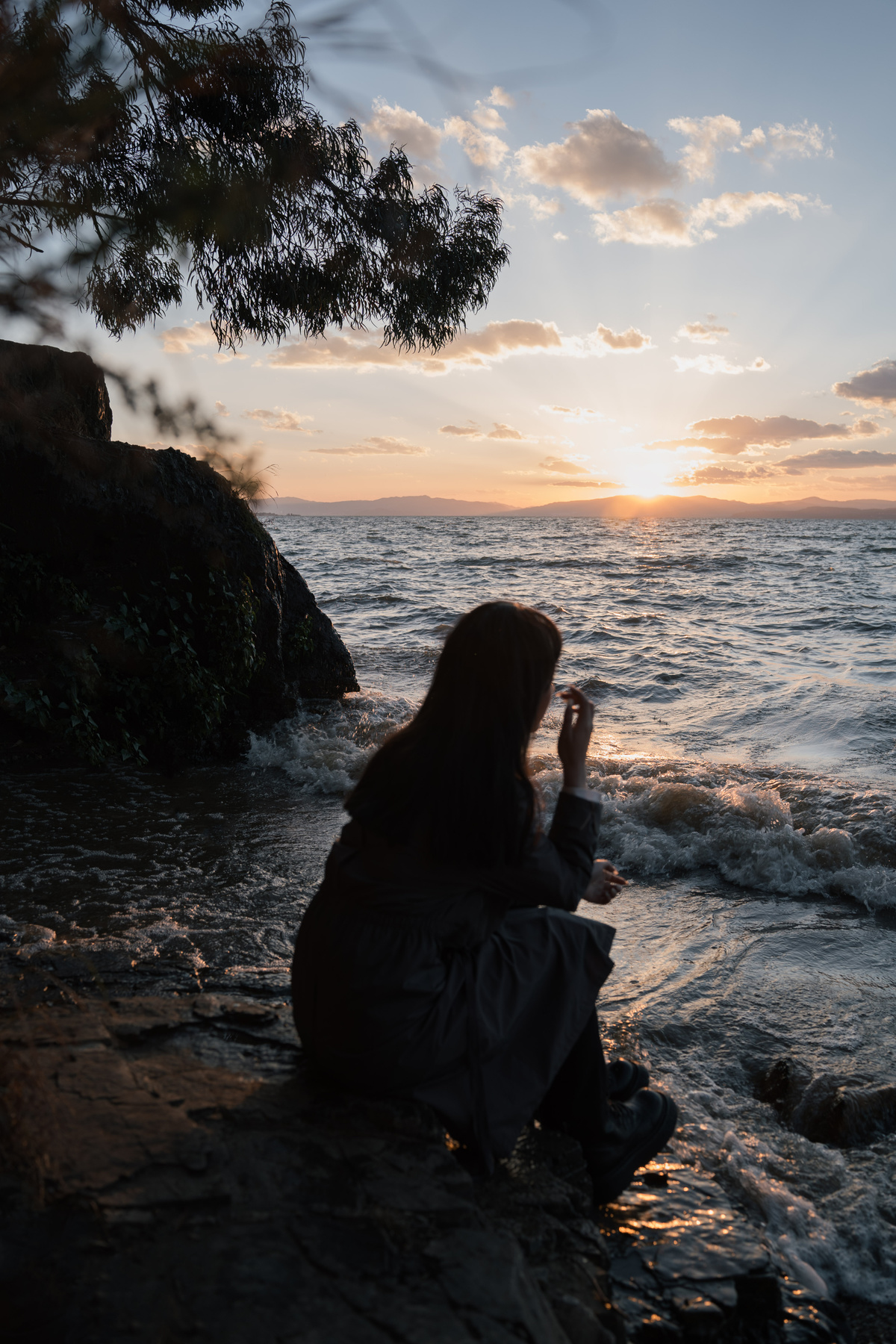 当太阳落山时 一名年轻女子坐在海边的岩石上 另一个人在拍照