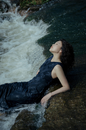 一位美丽的年轻女子穿着黑色连衣裙躺在河或小溪中的岩石上