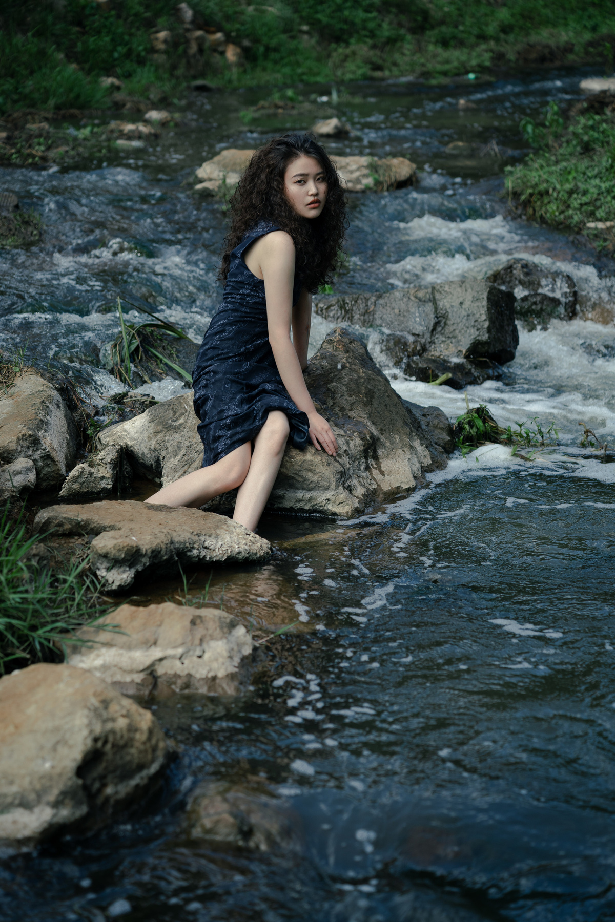 一个穿着黑色连衣裙的年轻女子坐在一条溪流中的岩石上