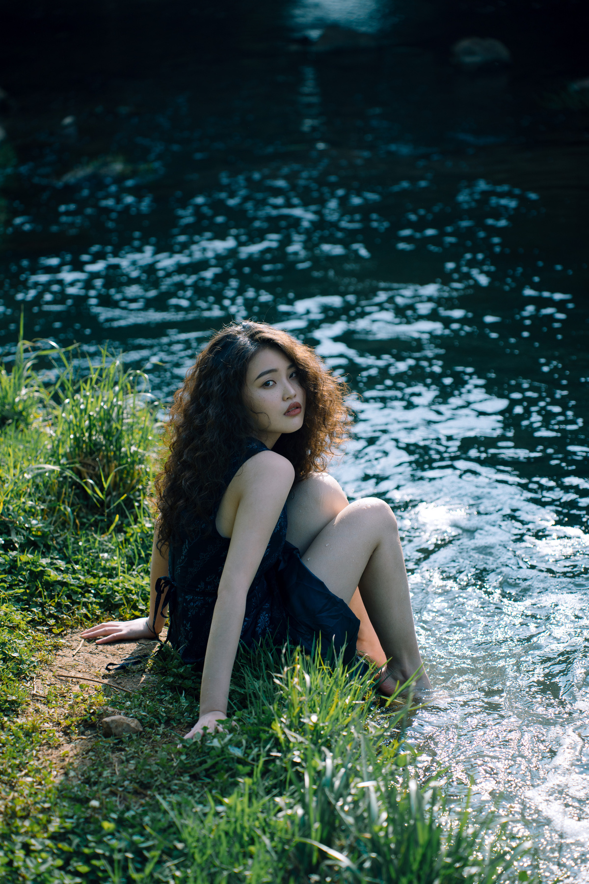 一个穿着黑色连衣裙的美丽年轻女子坐在河边或小溪旁的草地上