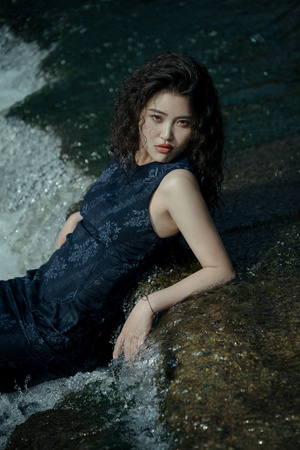 美丽的年轻女子穿着黑色连衣裙躺在河或小溪中的岩石上