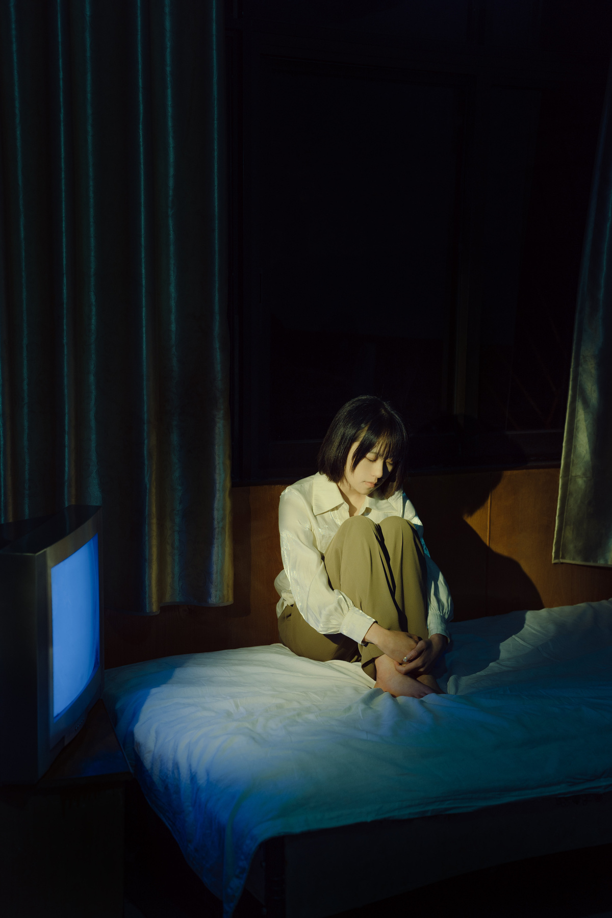 一个年轻女孩坐在一个黑暗的房间里 电视开着 另一个人也坐在床上。