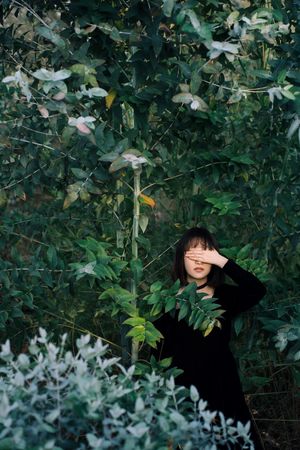 一个穿着黑色衣服的年轻女子站在一个长满灌木和树木的森林中 她的脸周围环绕着这些植物。