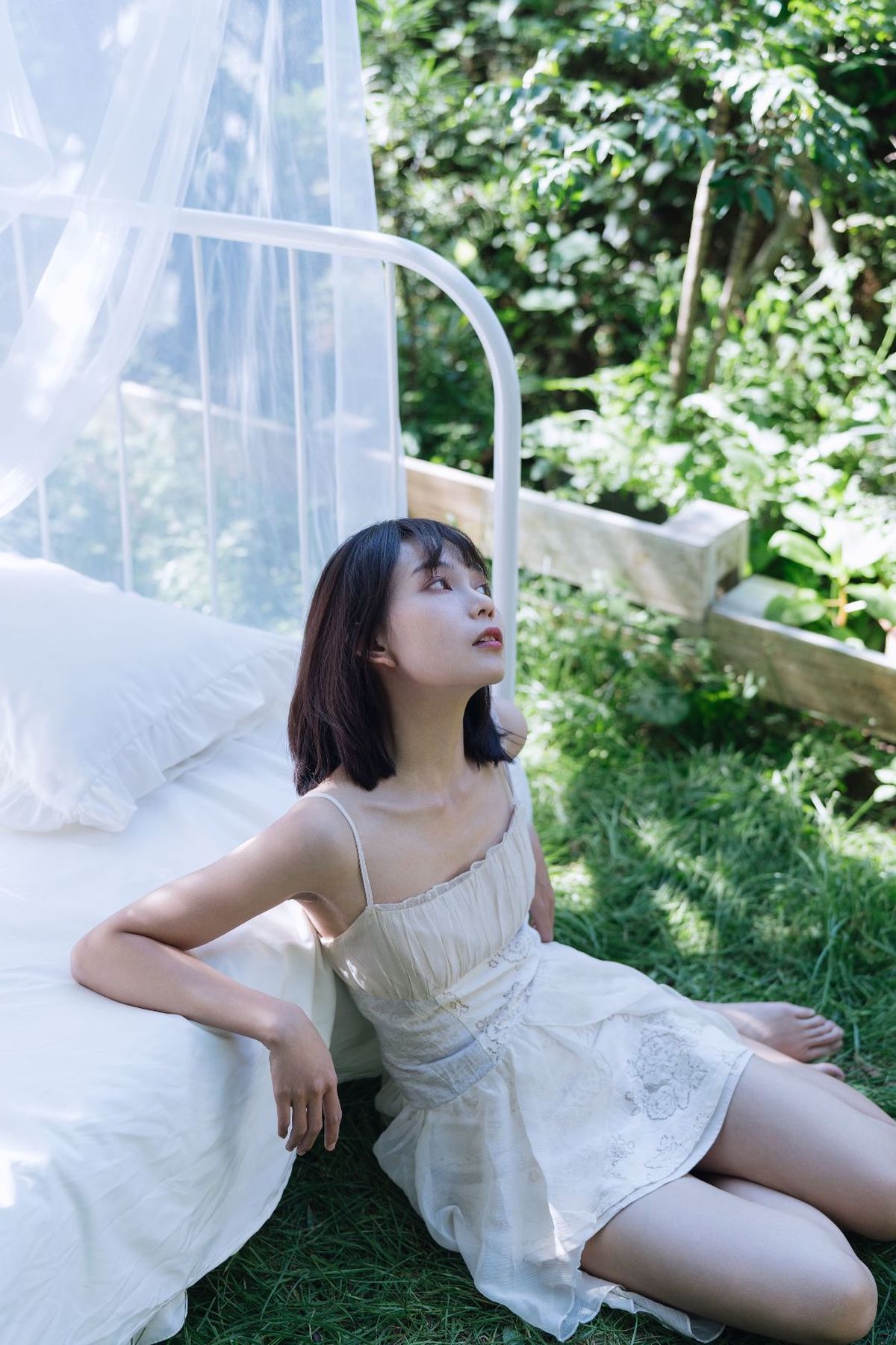 一个穿着白色连衣裙的年轻女子坐在床上 床单也是白色的 花园里。
