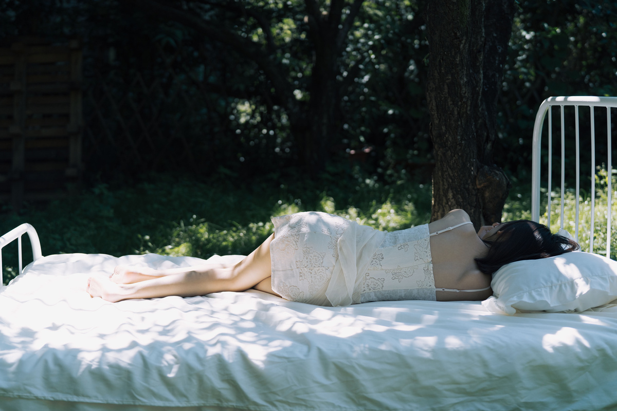 一位躺在床上的女人 旁边有白色的床单 旁边有一棵树。
