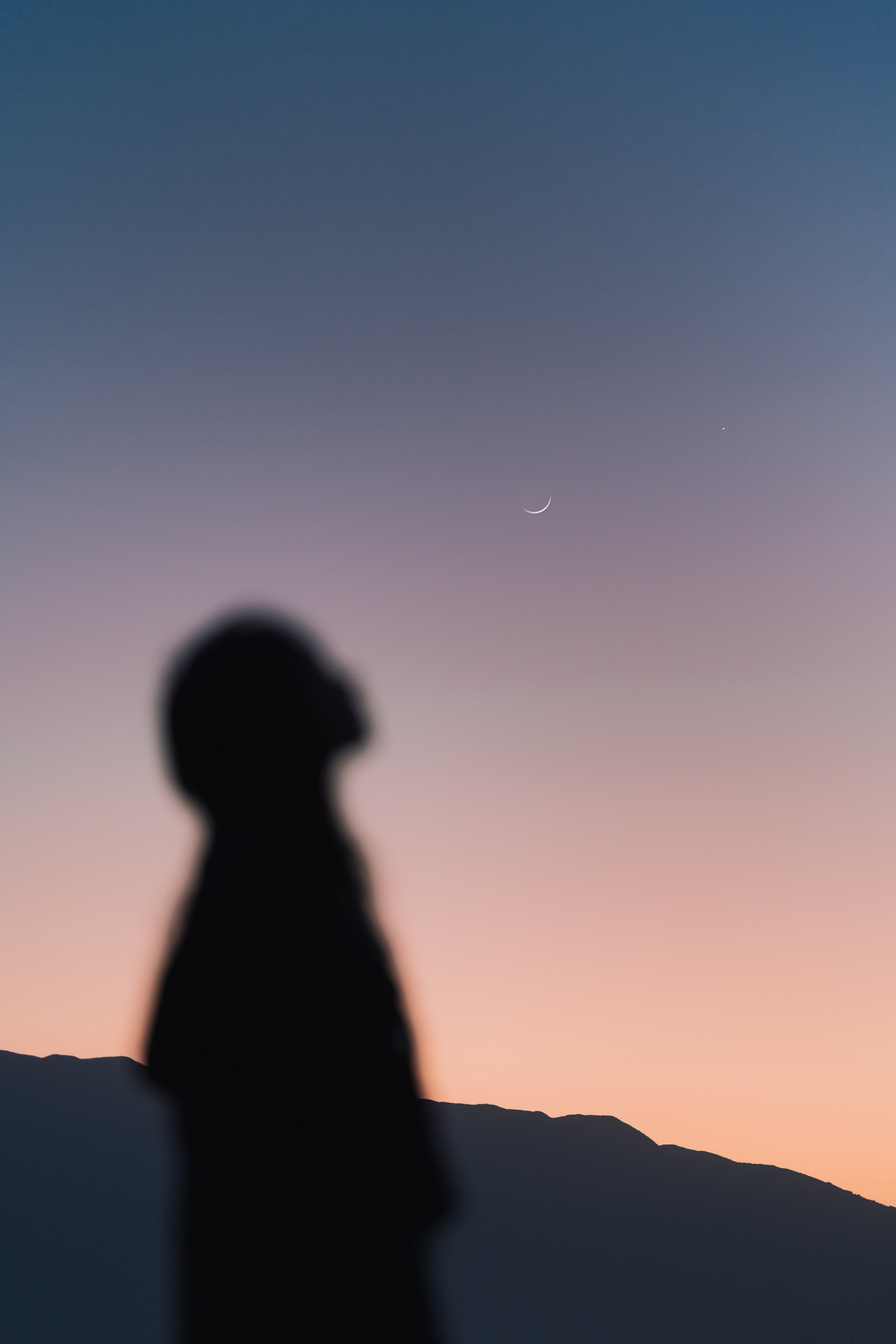 一个人站在山顶 看着日落 天空中挂着月亮的剪影。