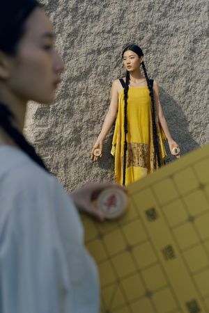 一位穿着黄裙子的年轻女孩站在一面墙前 一个男子在注视着她。