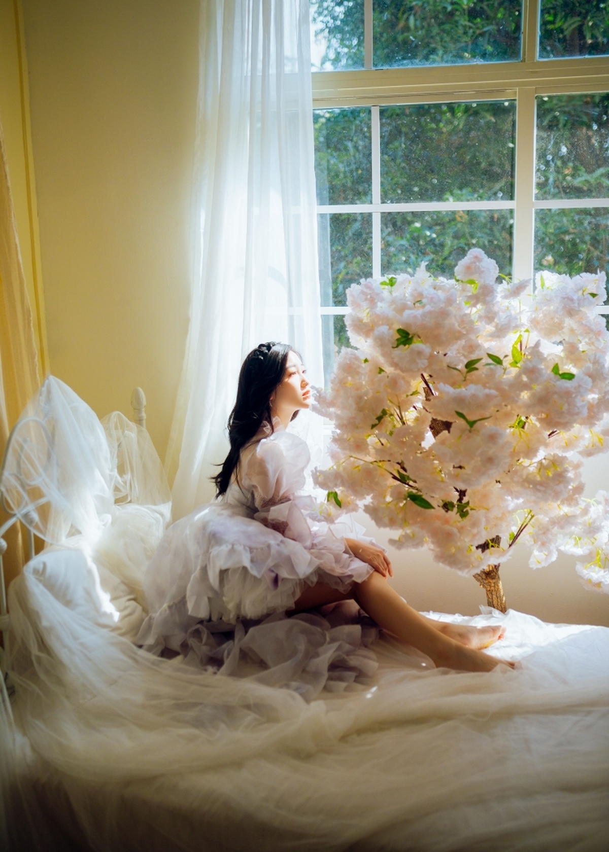 一个穿着白色连衣裙的年轻女子坐在窗前 窗外有一棵开满白色花朵的树。