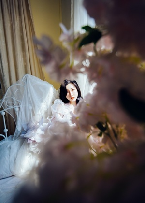一位穿着白色长裙和面纱的年轻女子正在窗前凝视一张床上盛开着鲜花的床。