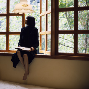 一个年轻女子坐在窗台上读书 向外看窗户外面的景色。