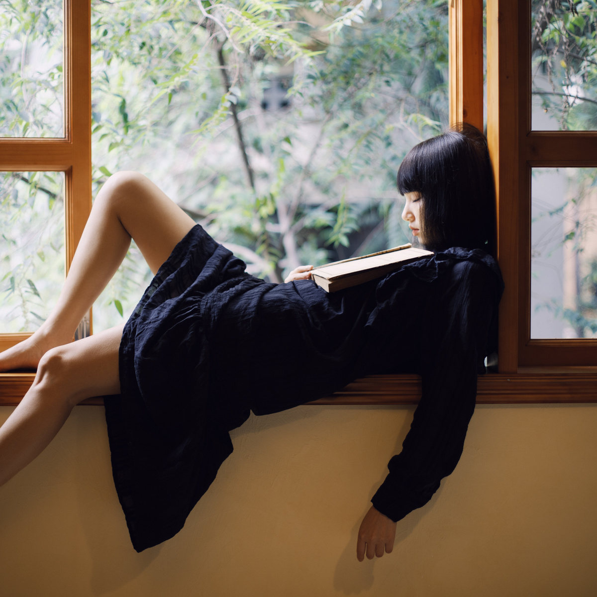 一个穿着黑色连衣裙的年轻女子坐在窗台上读书